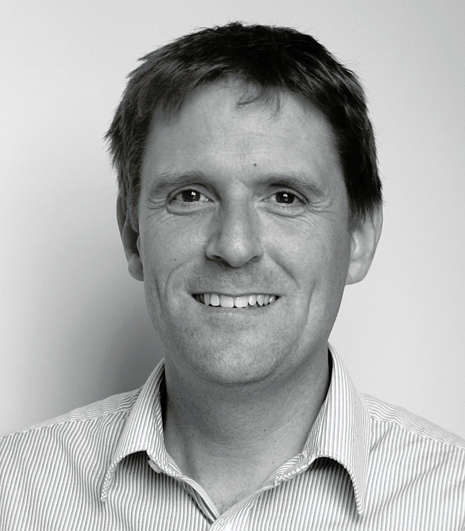 Black and white portrait of a man smiling, wearing a striped shirt with a neutral background.