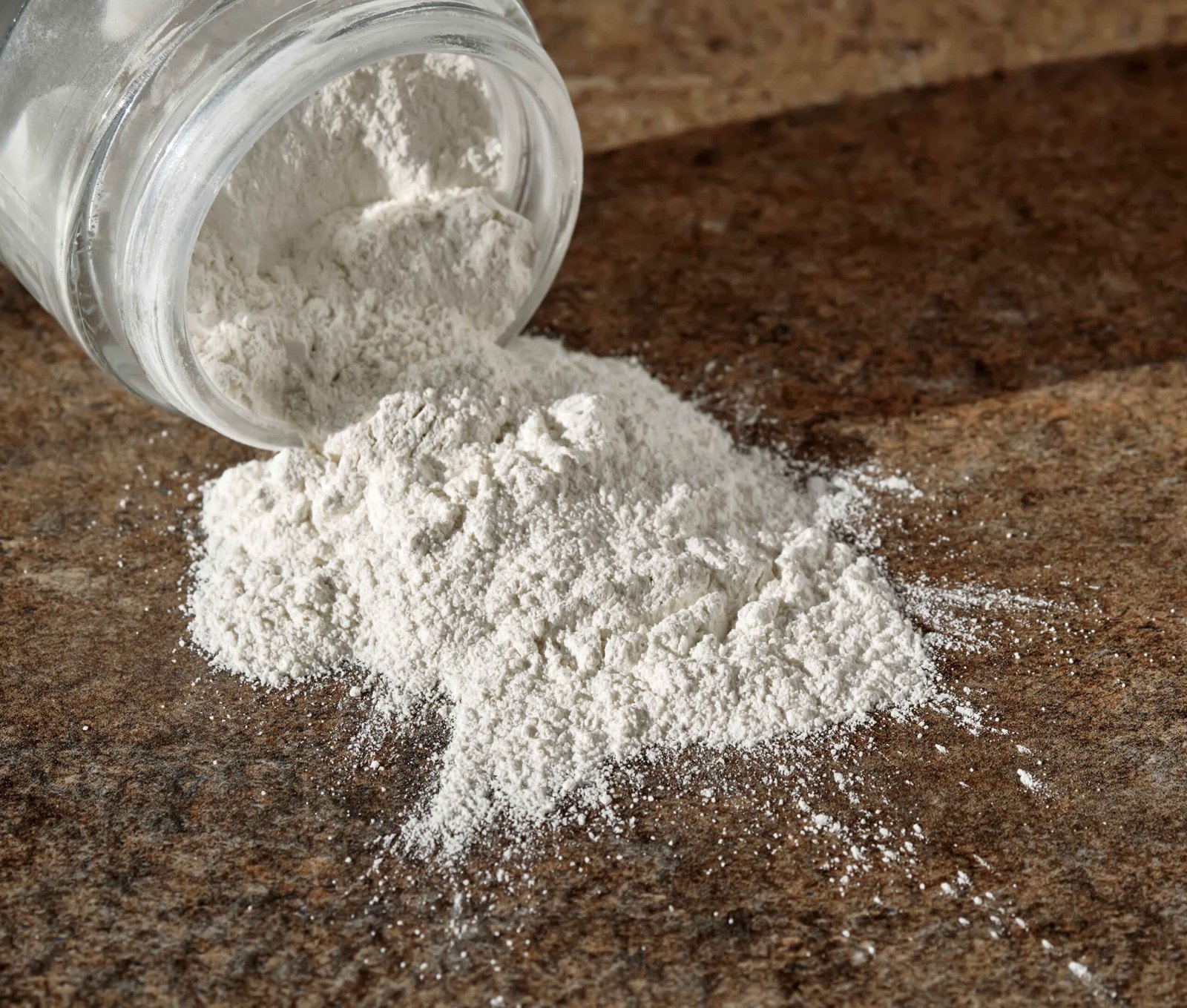 Glass jar tipped over with white powder spilling onto a surface, showing a fine texture and scattered appearance.