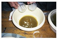 Hands pouring liquid ingredients into a large bowl containing a brown mixture on a wooden surface.