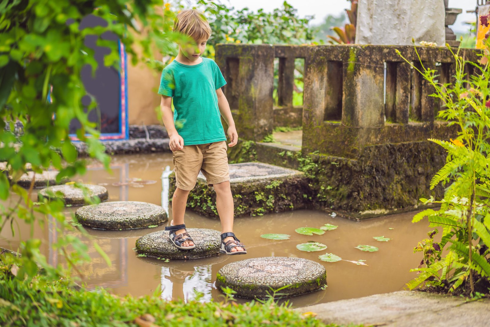 Young boy in a green t-shirt walking on circular stepping stones over a pond with lily pads.