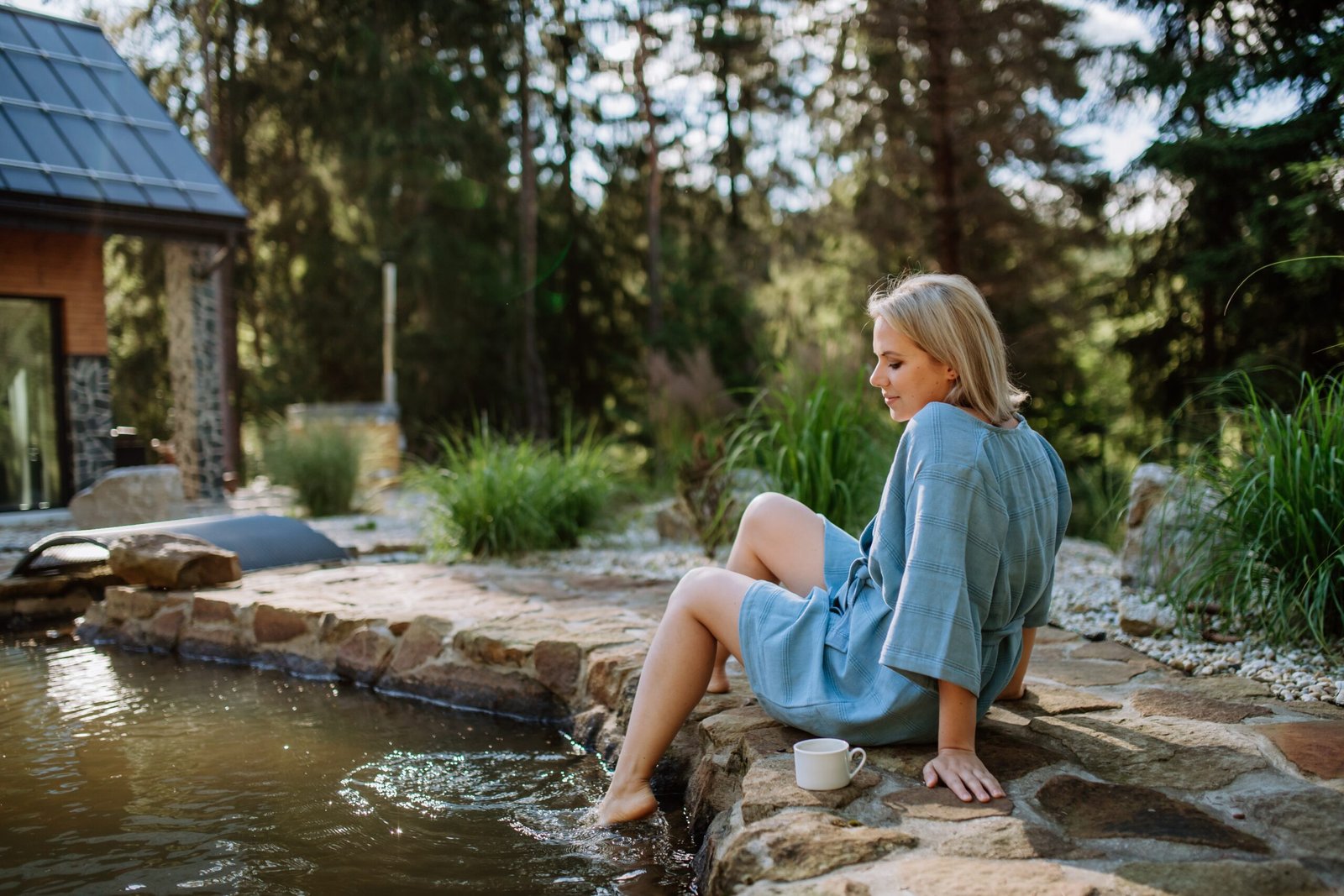 Woman in a light blue robe sitting on the edge of a stone pond with a cup in hand, enjoying the natural surroundings.