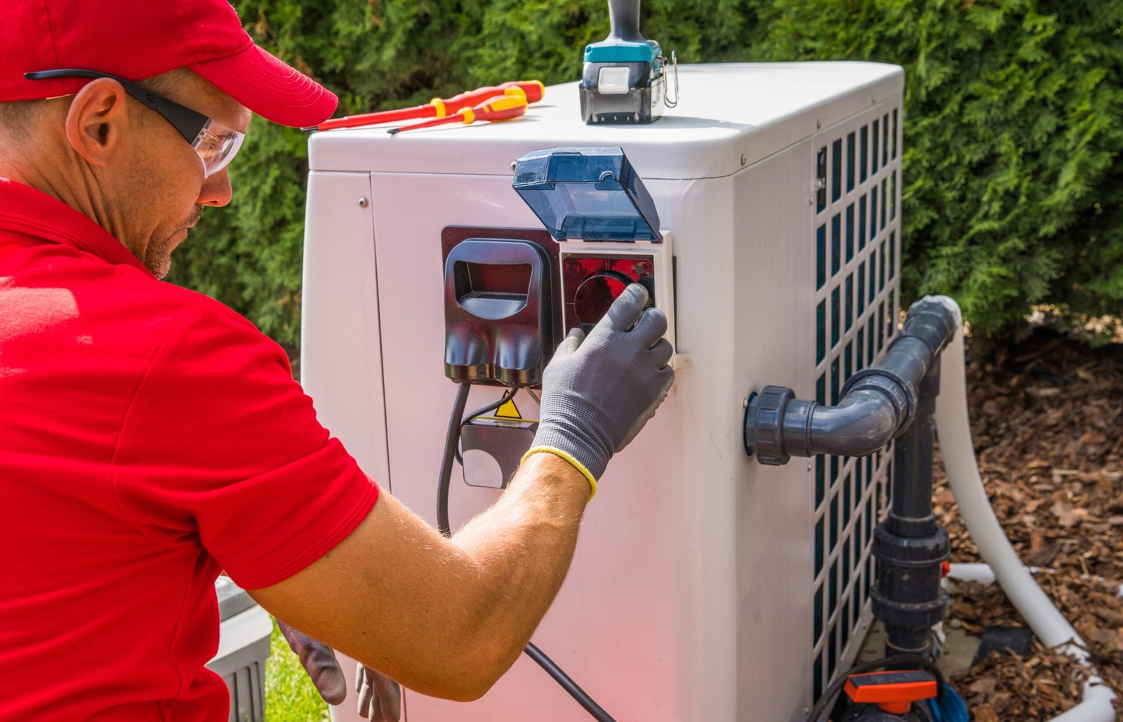 Technician in red shirt adjusting the control panel on a pool heater unit outdoors.