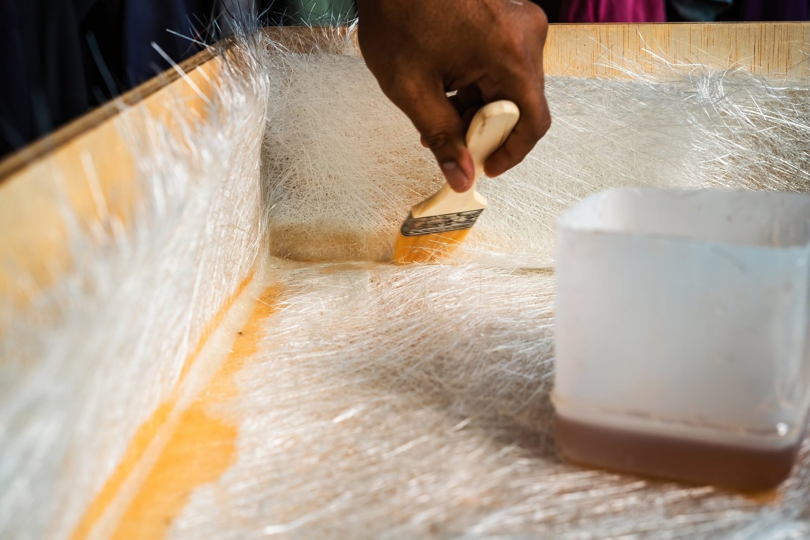 Person using a brush to apply resin on fibreglass strands in a wooden container.