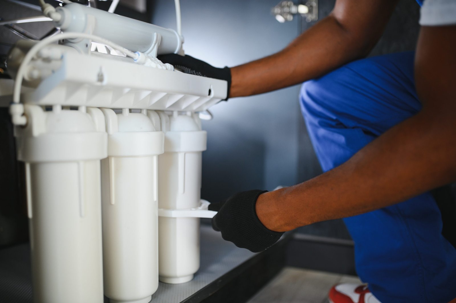 Person in blue overalls installing a water filtration system under a sink with white filter cartridges.
