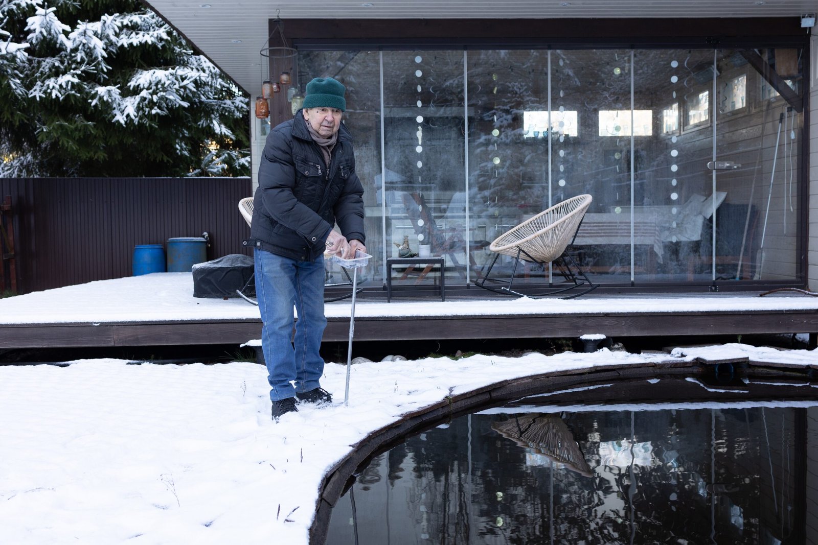 Man in winter clothing standing beside a pond with snow on the ground and a wooden deck in the background.
